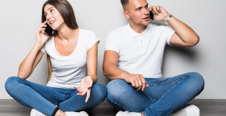 Young couple talking on the phone while sitting on the floor isolated on gray background