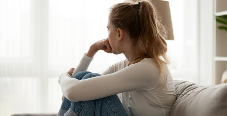 Side view young woman looking away at window sitting on couch at home. Frustrated confused female feels unhappy problem in personal life quarrel break up with boyfriend or unexpected pregnancy concept