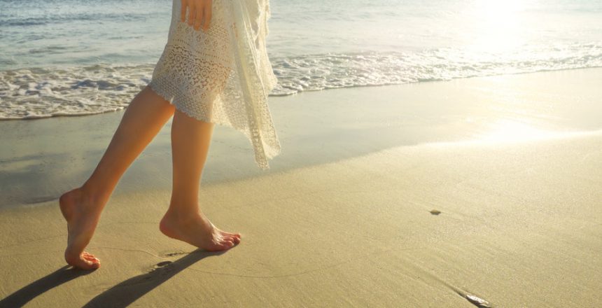 Young girl in white dress walking alone on the sandy beach at sunrise.
Closeup detail of female feet and golden sand on beach.